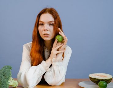 Woman with red hair holding lime surrounded by fresh produce on table.